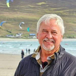 Alfred O’Neill smiling on a beach, with kite surfers and green hills behind him.